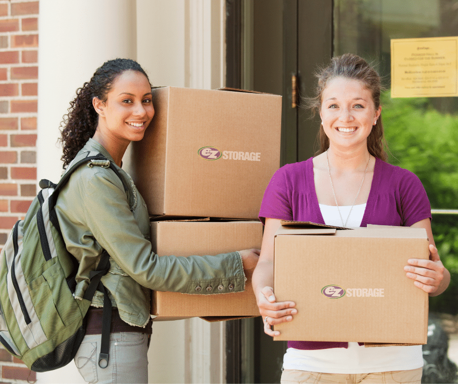 Two women standing outside a college dorm holding moving boxes.