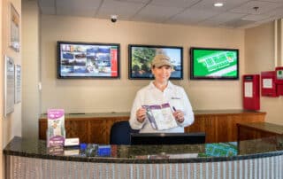 Self storage manager at EZ Storage holding a brochure, standing behind a counter with security surveillance monitors in the background..