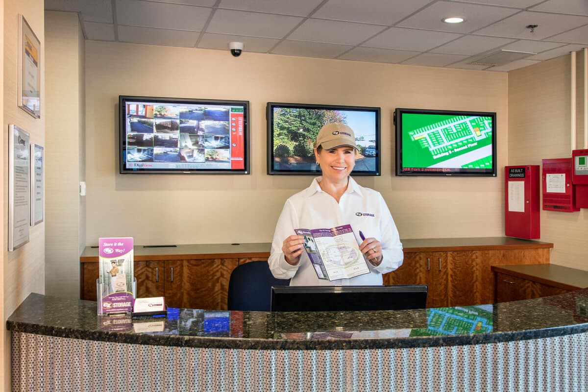 Self storage manager at EZ Storage holding a brochure, standing behind a counter with security surveillance monitors in the background..