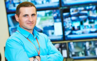 man wearing a security badge standing in front a group of security surveillance displays