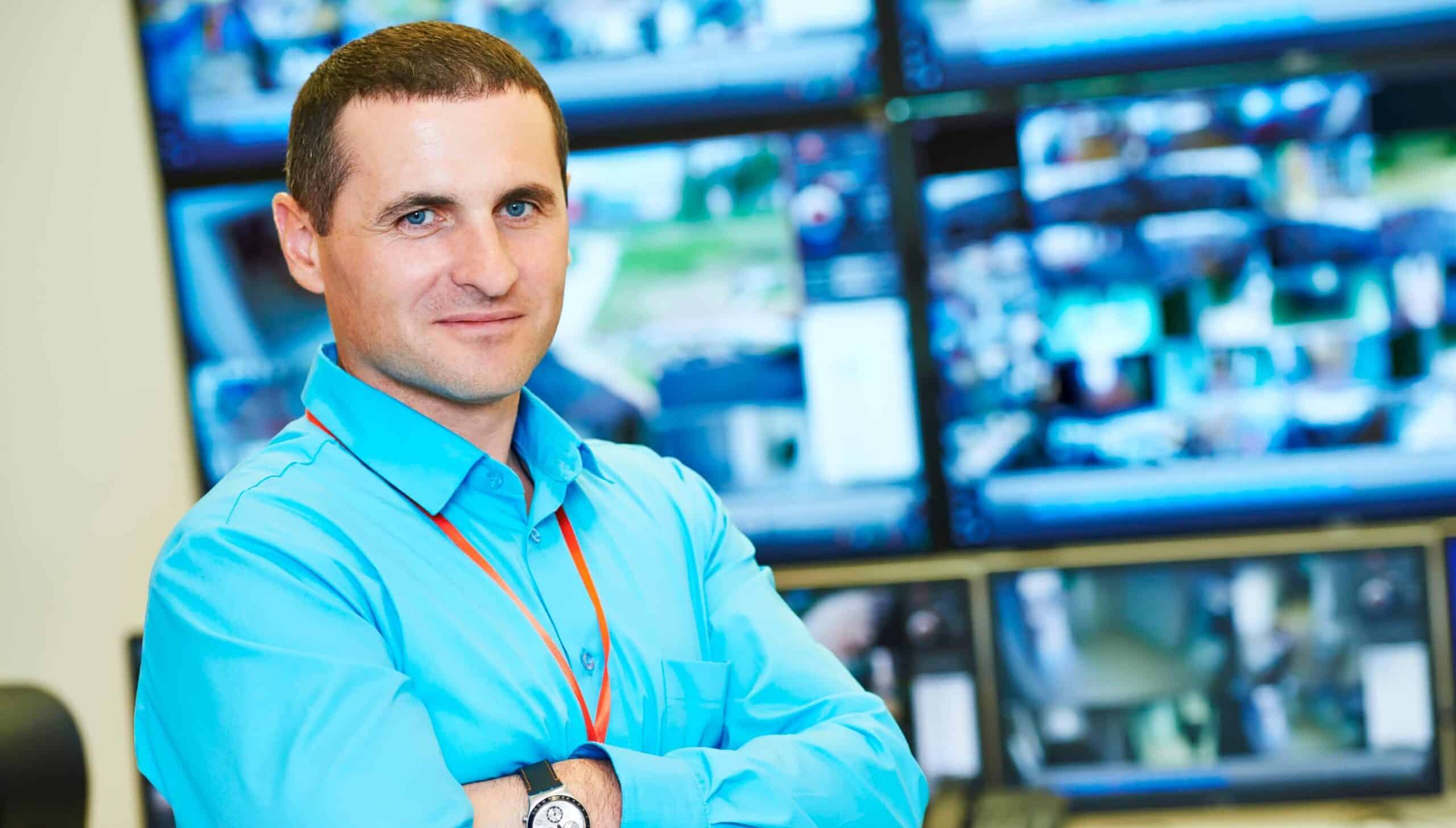 man wearing a security badge standing in front a group of security surveillance displays