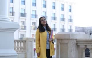 young woman in a yellow coat holding text books, standing outside a concrete building