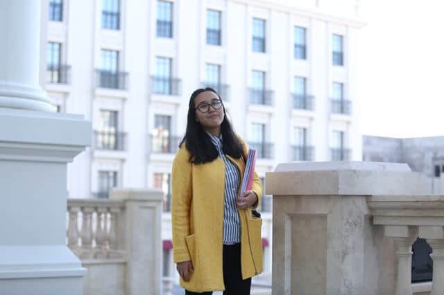 young woman in a yellow coat holding text books, standing outside a concrete building